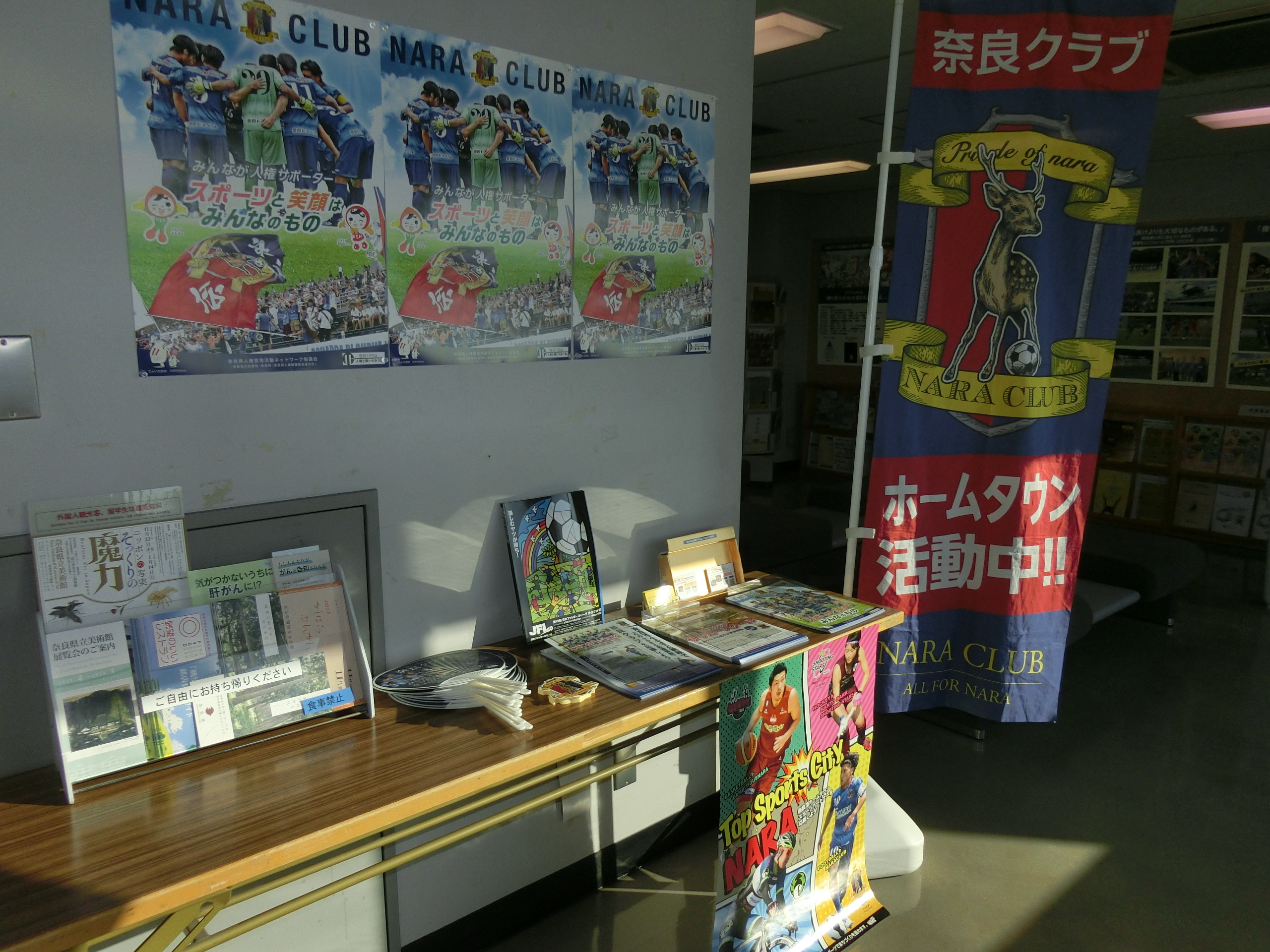 ホームタウン活動中と書かれたのぼりと、うちわ・ステッカー・パンフレット等のグッズ